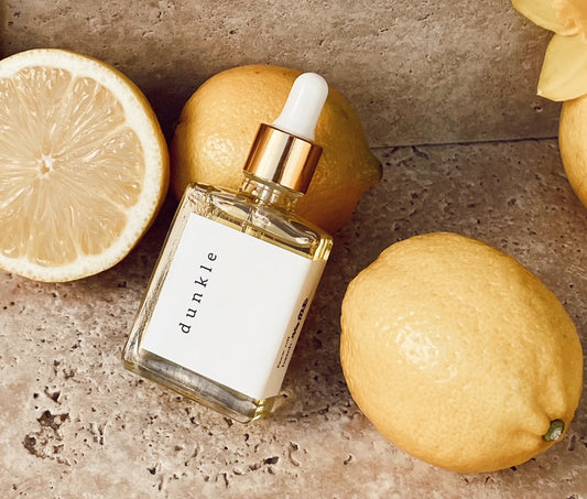 Dunkle face oil bottle sitting on beige travertine with yellow daffodil flowers popping into frame at the forefront.