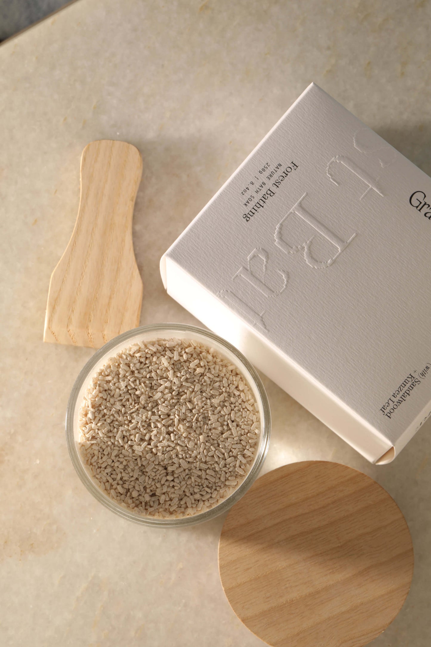 An open jar of Gracious Minds Forest Bathing Soak. Next to the soak is a timber scoop and lid, as well as the white Gracious Minds product box.