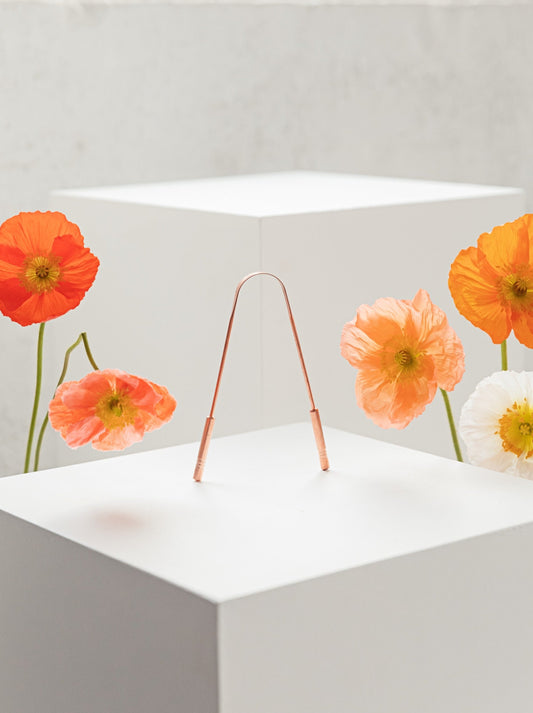 Copper tongue cleaner balancing on a white plinth with fresh poppies either side