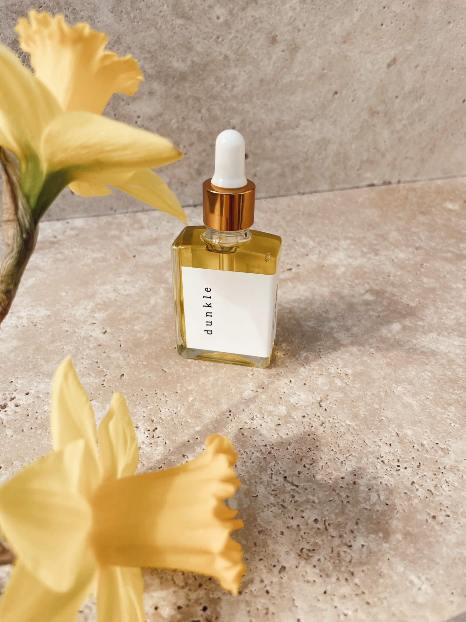 A bottle of Dunkle facial oil styled with Self Bloom yellow daffodils on a stone bathroom benchtop.