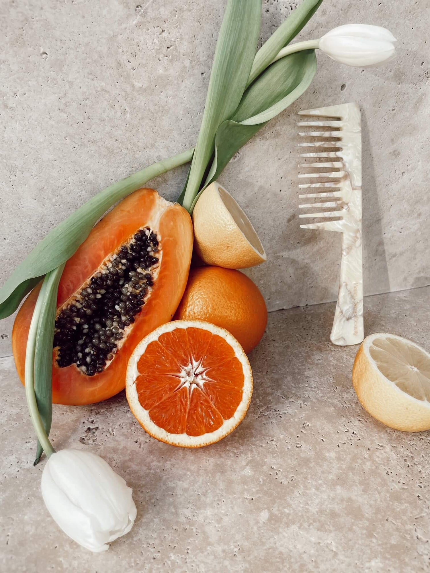 Base Camp Beauty buttermilk comb leaning on a stone bathromm benchtop amongst papaya, oranges, lemons and white tulips.