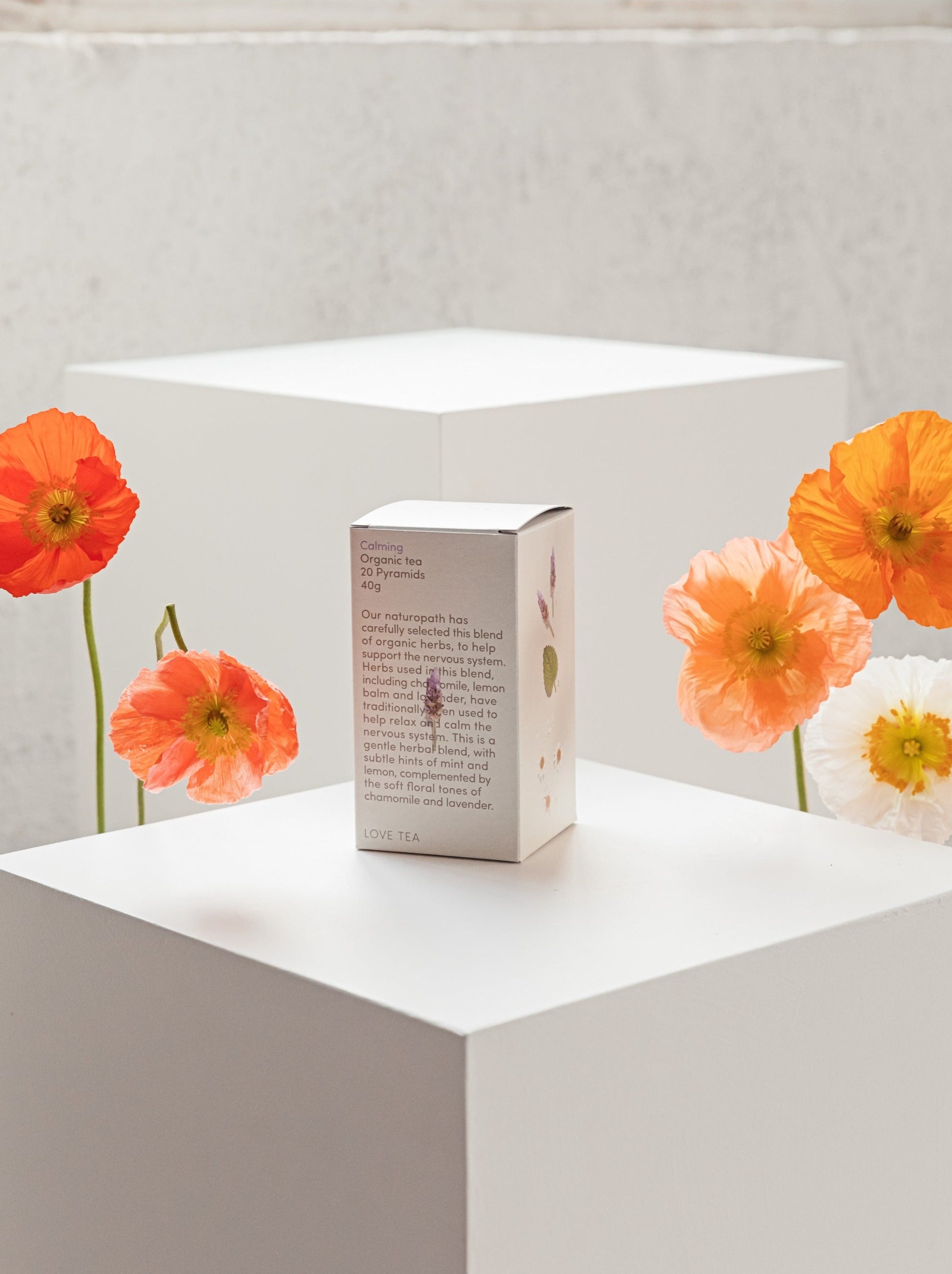 Box of tea sitting on a white plinth with fresh poppies on each side of the product