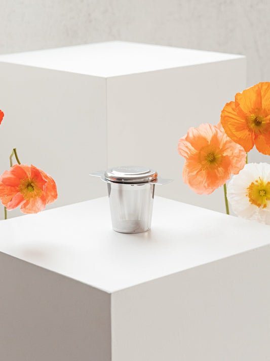 A silver steel tea infuser with metal lid sitting on a white plinth with fresh poppies on each side of the product
